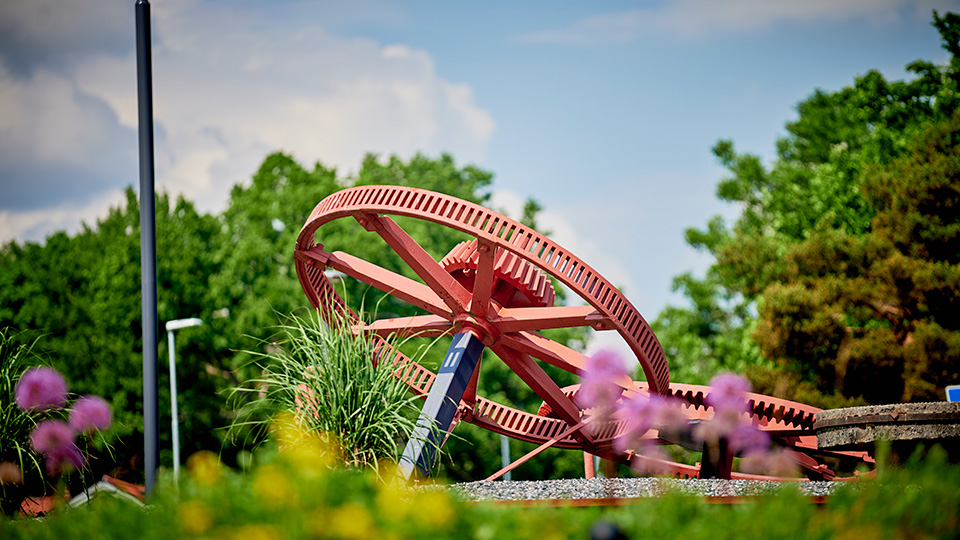 Ett stort, rödmålat kugghjul står på en grönskande gräsplätt.