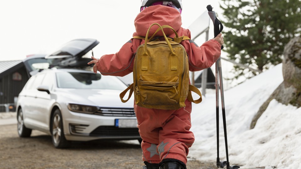 Ett barn promenerar iväg mot en bil med vinteroverall, ryggsäck och stavar i handen. 