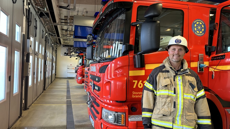 Bilden visar en brandman i full utstyrsel framför en brandbil i ett garage på brandstationen