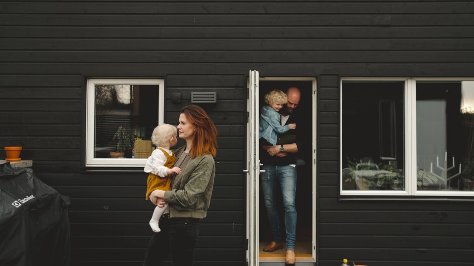 En mamma bär på sin bebis och lämnar huset medan pappan står kvar i dörröppningen med ett äldre barn. 