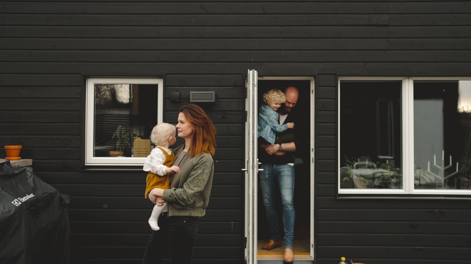 En mamma bär på sin bebis och lämnar huset medan pappan står kvar i dörröppningen med ett äldre barn.
