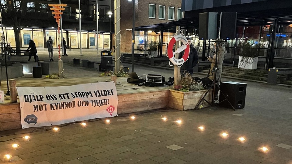 Ett foto taget på kvällen när det blivit mörkt vid Storängstorget. Marschaller brinner i en ring och ett plakat med texten Hjälp oss att stoppa våldet mot kvinnor syns i bakgrunden.