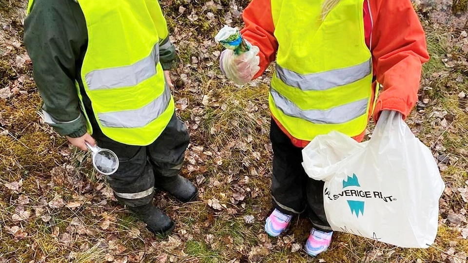 Två galonklädda barn står bredvid varandra utomhus. På marken ligger en massa döda löv och i handen håller det ena barnet en plastpåse där det står Håll Sverige rent