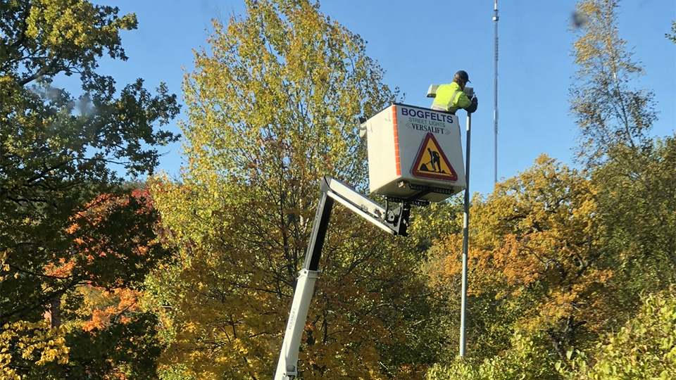 En man i gula varselkläder står i en korg till en skylift och inspekterar en gatlykta.