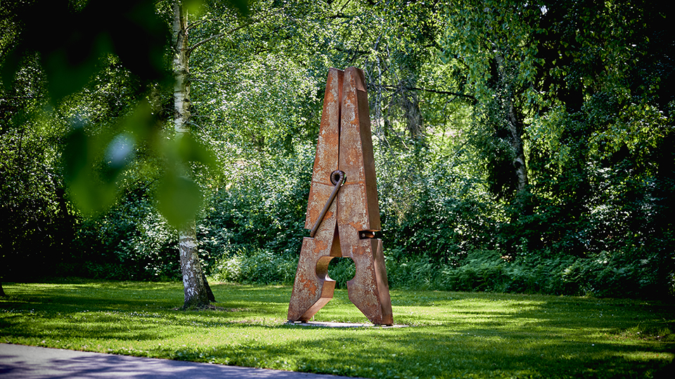 En gigantisk klädnypa i rostigt järn står upprätt under skuggan av träd.