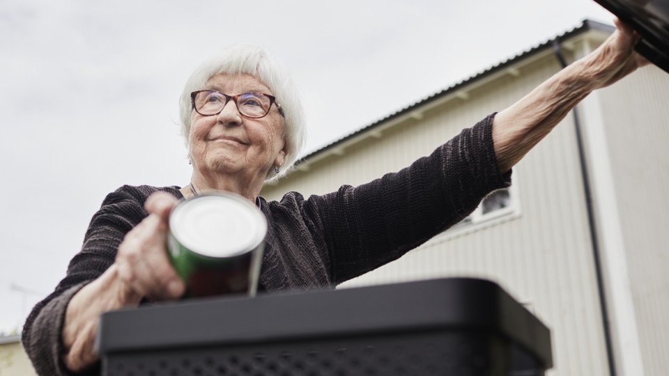 Äldre kvinna håller i en tom konservburk i ena handen och ett öppet lock till sopkärl i den andra handen.