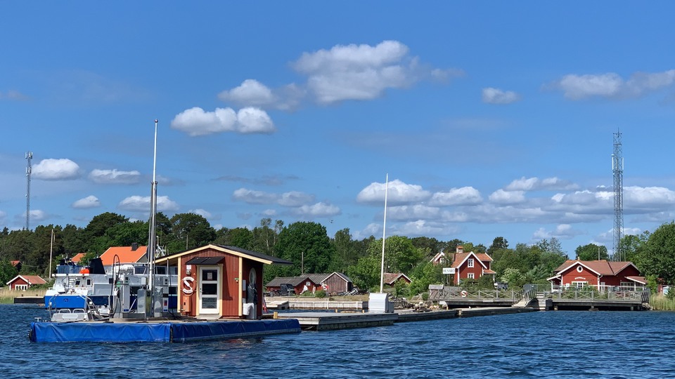 Brygga på Husarö med bränslepump, en sjömack, och röda hus i bakgrunden.