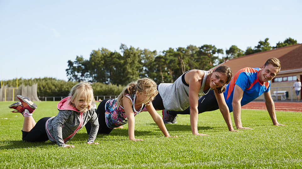 Familj med två vuxna och två barn tränar i gräset på en idrottsplan.