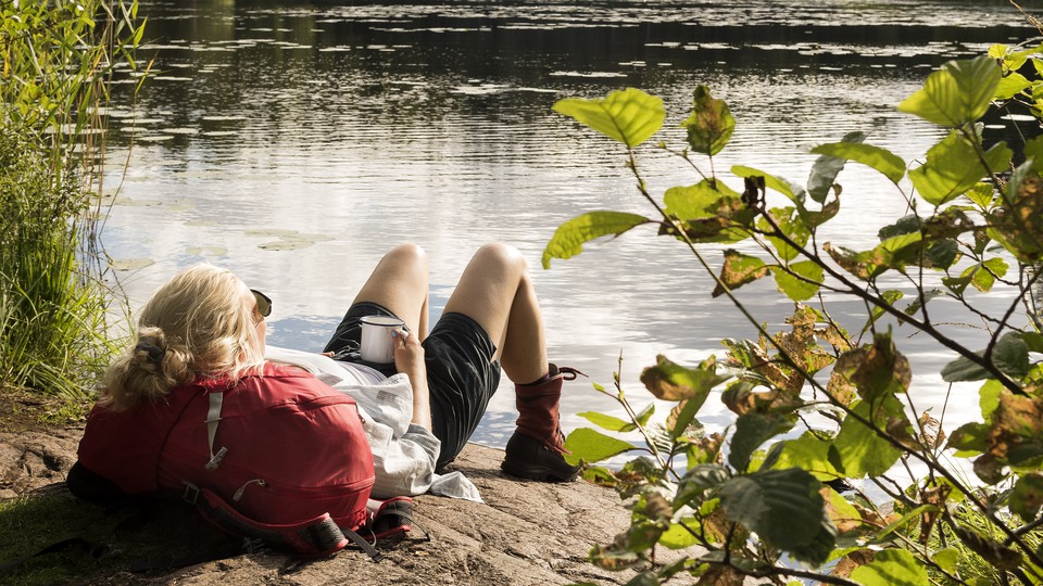 En kvinna med ryggsäck ligger och vilar på en klipphäll med sjön Drängsjön i bakgrunden. Solen skiner och det ser varmt ut.