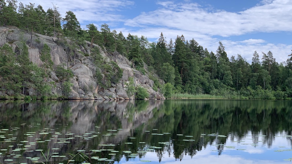 En stilla sjö där skog och berg speglar sig i vattnet. 