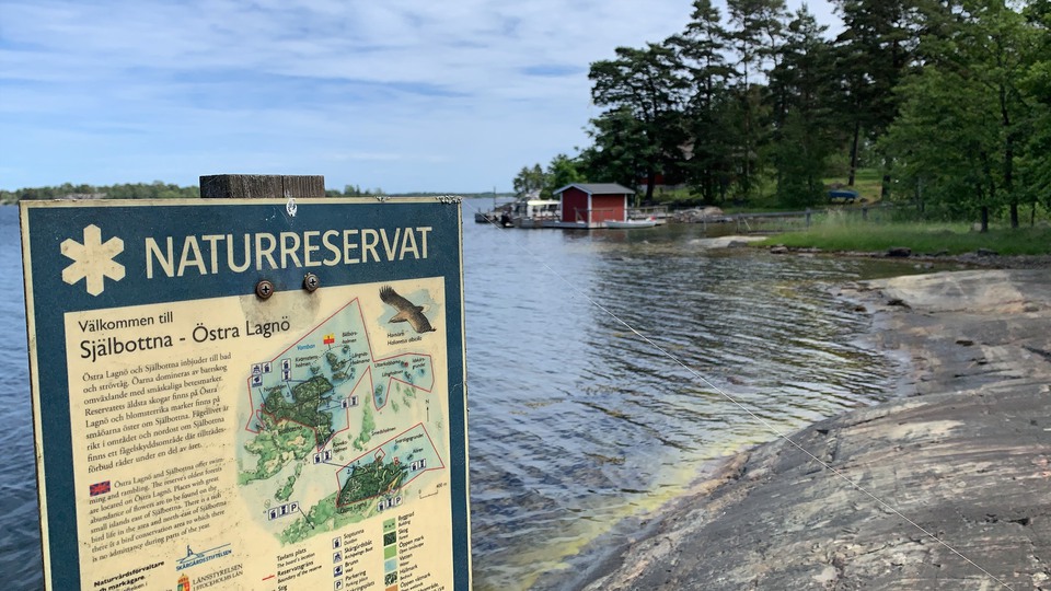 En skylt som visar information om naturreservatet Själbottna - Östra Lagnö. Skylten står vid en havsvik med klipphällar.