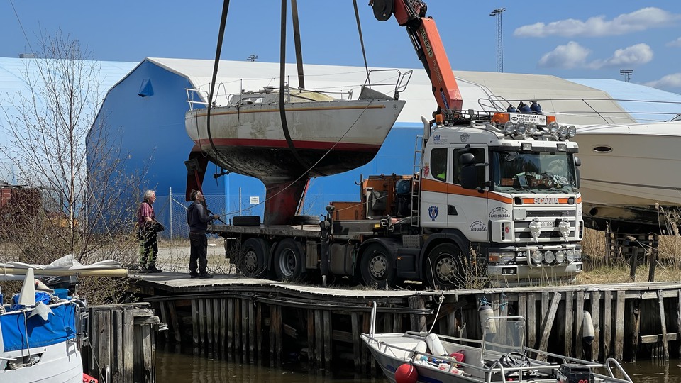 En sliten segelbåt lyfts upp ur vattnet med hjälp av en lyftkran på en stor lastbil. Två personer står bredvid och styr kranen.