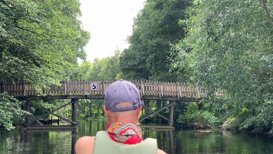 Du ser en person bakifrån som paddlar under en välvd träbro över kanalen.