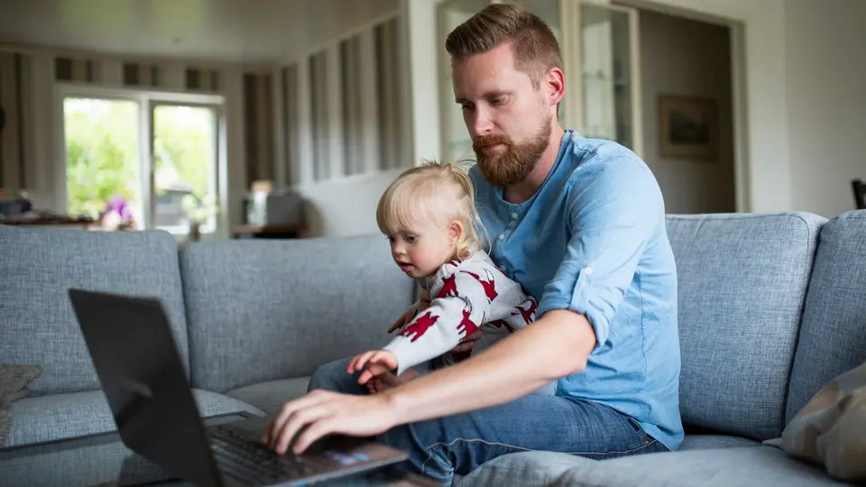 En man sitter i en grå soffa i ett större vardagsrum. I sin famn har han ett litet barn och framför honom finns en dator på ett soffbord. 
