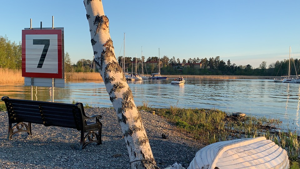 En tom parkbänk längst ut på en udde med utsikt över vatten och segelbåtar som ligger förtöjda vid bryggor.