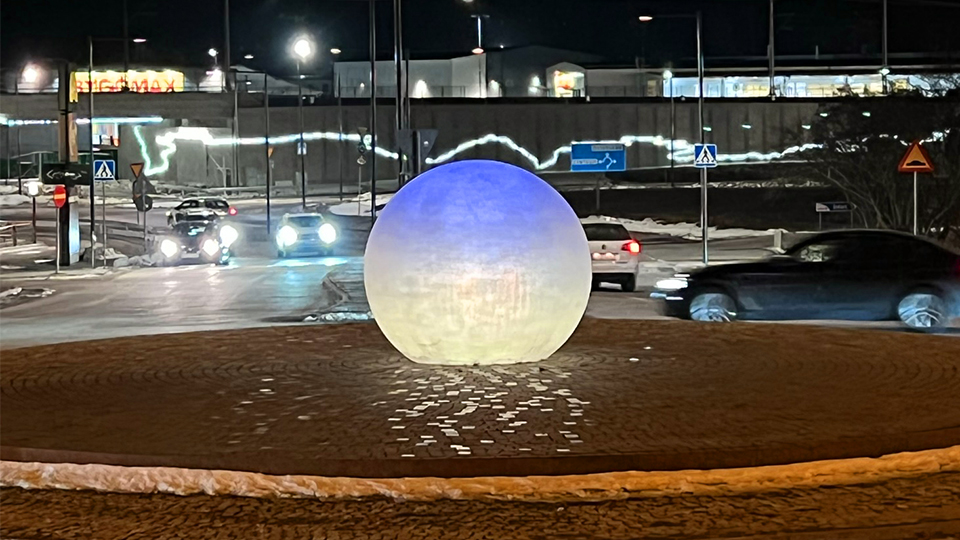 En rondell på kvällen. Det är mörkt, men solen (en slags boll) mitt i rondellen är upplyst så att den undre delen lyser gult och den övre delen lyser blått.