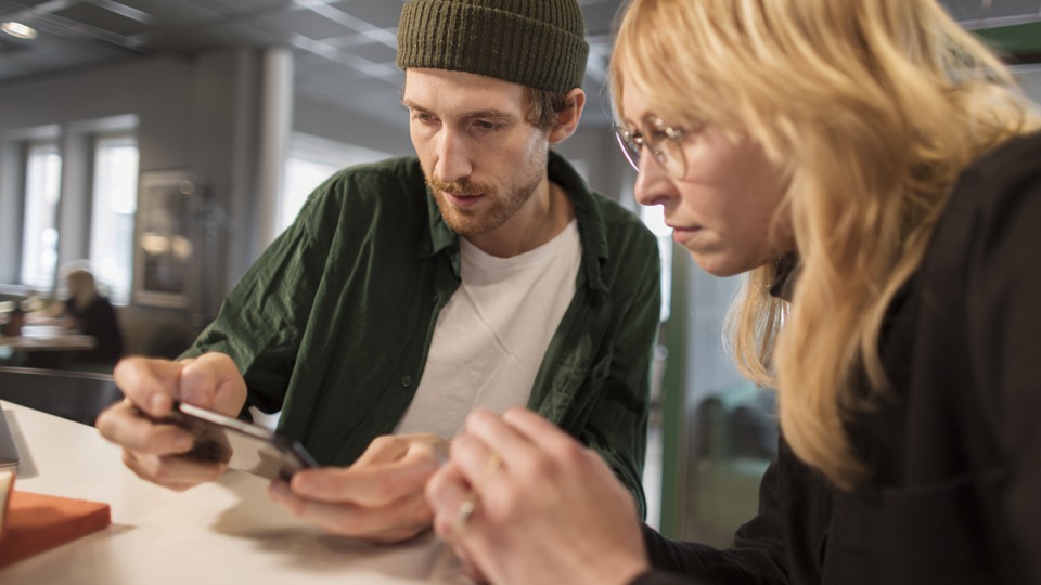 Två personer sitter på samma sida ett bord och tittar på en mobilskärm