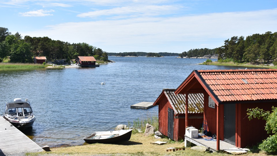 Faluröda sjöbodar vid en stilla vik. En brygga med en motorbåt. En roddbåt ligger på land. 