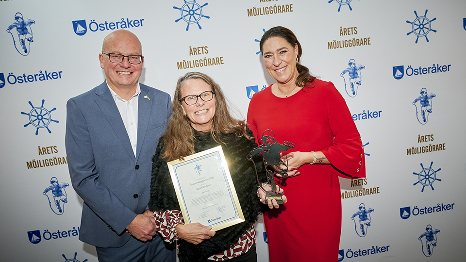 Årets möjliggörare 2022, Karin Olofsson står tillsammans med kommundirektör Staffan Erlandsson och Kommunstyrelsens ordförande Michaela Fletcher (M) vid en fotovägg med Österåkers logotyp, texten Årets möjliggörare och en stiliserad bild av statyetten Level up av Tomas Lacke.