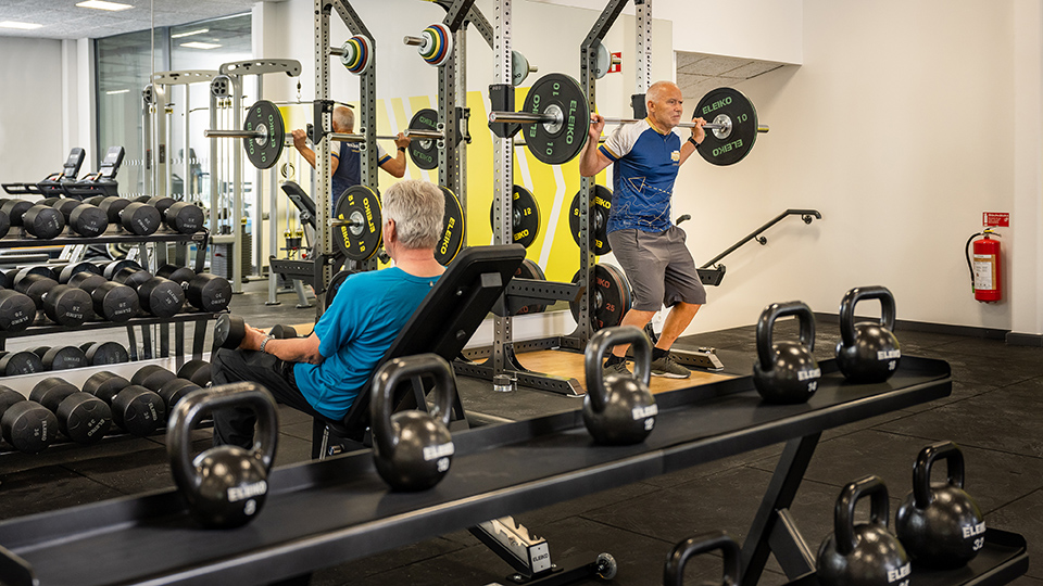 Bilden visar två män som tränar i gymmet. Redskap som syns i bild är kettlebells, skivstång, spegel och fria vikter.