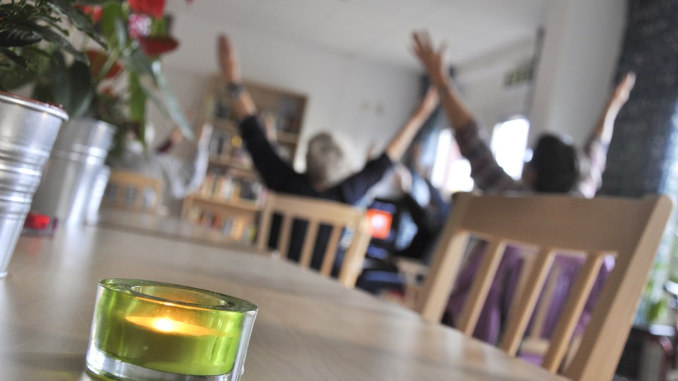 En grupp äldre utför en aktivitet. De sitter ner och sträcker upp armarna i luften. I bilden syns även ett dukat bord med en blomma och ett tänt ljus.