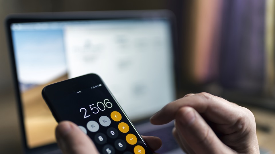 Bilden föreställer händer som använder en kalkylator i en mobiltelefon framför en laptop.