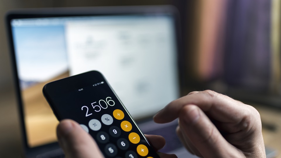 En person håller i en telefon som visar en miniräknare. I bakgrunden står en dator.