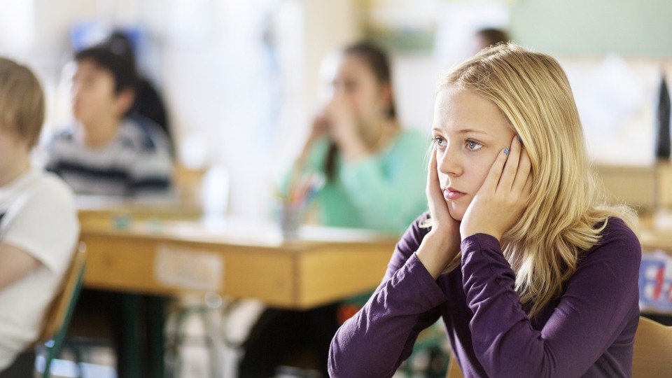 Flicka sitter i bänken i klassrum. Hon ser allvarlig ut. Har huvudet i händerna. Runtomkring henne sitter andra elever, det pågår en lektion.