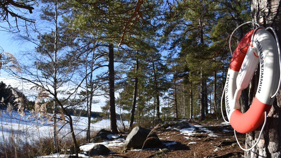 I förgrunden till höger i bild hänger en livboj på en ställning. I mitten av bilden är strandkanten med några stora stenar och till vänster is- och snötäckt sjö. stig
