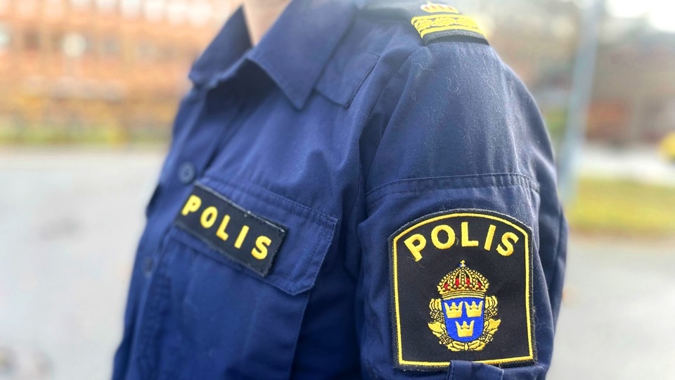 Bilden visar vänster sida på en polisuniform. Uniformen är mörkblå och på ärmen sitter ett påsytt emplem som det står Polis på. I bakgrunden syns Alceahusets rödbruna tegelvägg.