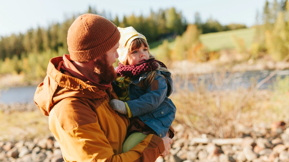 En pappa håller i sitt barn i famnen. De står på en strand och har ytterkläder på sig