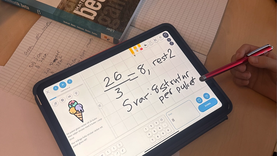 En surfplatta ligger på en skolbänk tillsammans med en matematikbok. På surfplattan syns det digitala verktyget i matematik som heter Magma.