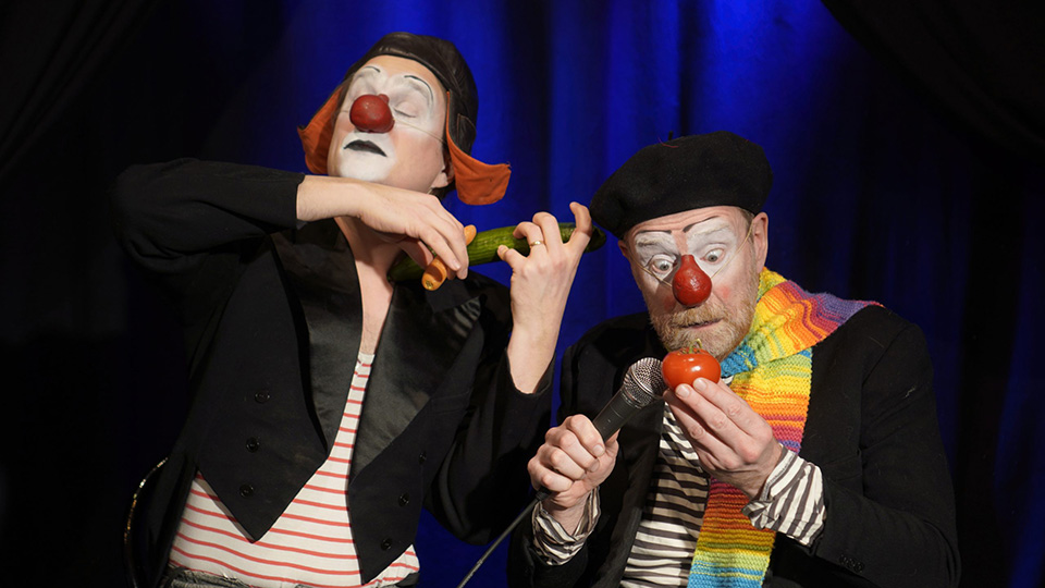 Två clowner med röda näsor. Den ena spelar på en gurka som om den var en fiol och den andra tittar på en tomat.