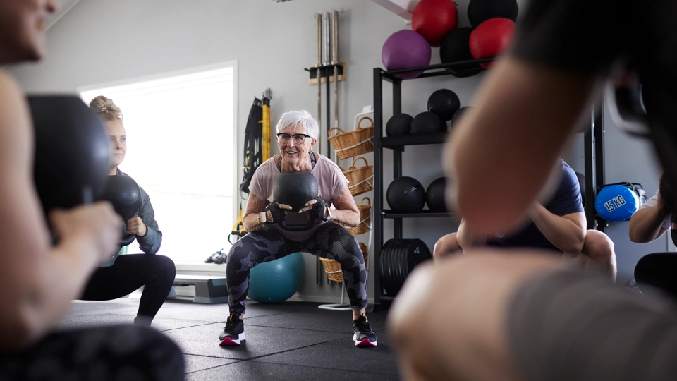 Äldre kvinna tränar på gym tillsammans med andra personer.