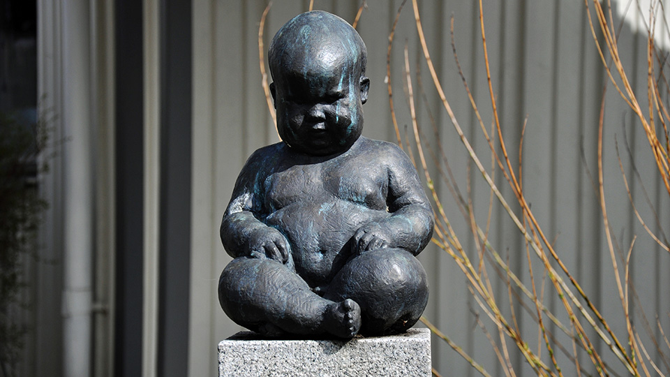 En knubbig bebis i brons. Hen sitter med benen i kors och med händerna vilande på knäna.