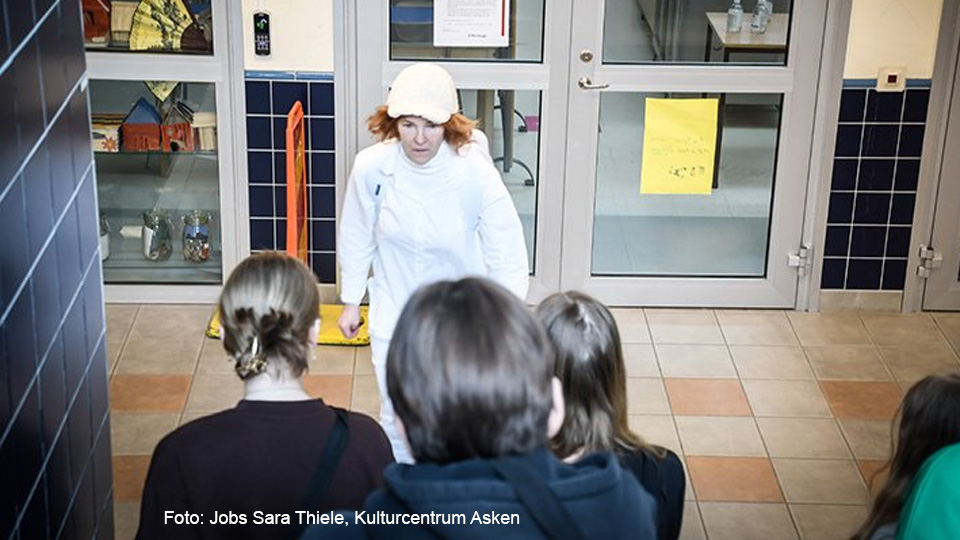 En helt klädd i vitt talar till en grupp ungdomar med ryggen mot kameran. De ser ut att sitta i en skolkorridor.