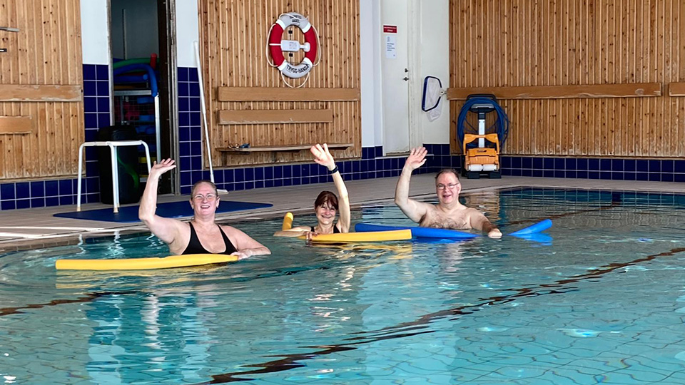 Tre personer i badkläder står i en bassäng och vinkar mot kameran.