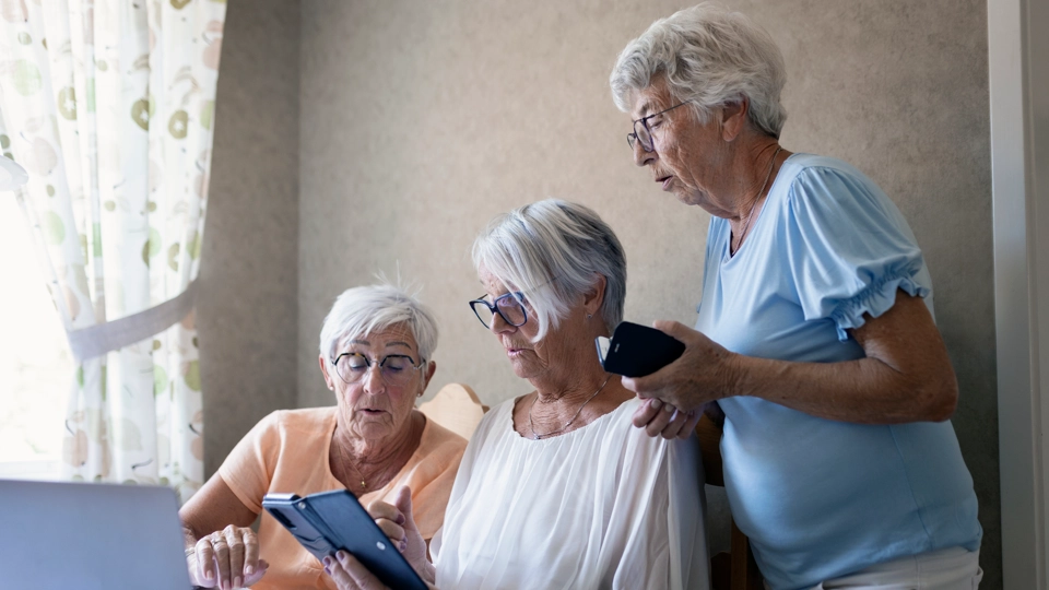 Tre äldre personer sitter tillsammans vid ett bord och tittar på en surfplatta och en mobiltelefon.