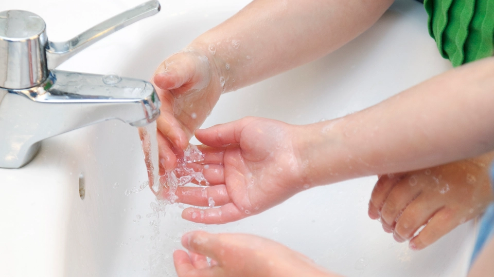 En handfat. Kranen rinner. Två barn tvättar sina händer.