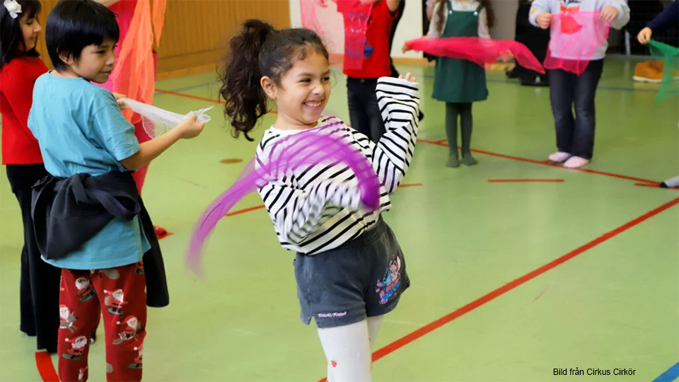 Glada barn i en gymnastiksal. De viftar med färglada schiffonsjalar.