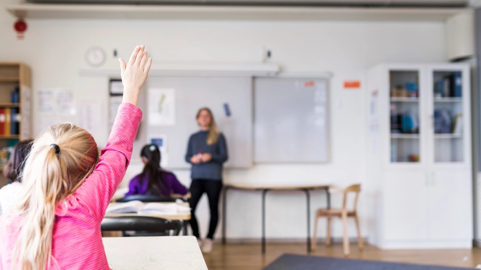 En lärare står längst fram i klassrummet. En flicka sitter vid en skolbänk och räcker upp handen.