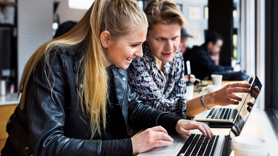 Två personer sitter med varsin bärbar dator på något som ser ut att kunna vara ett café. De samtalar engagerat med varandra.