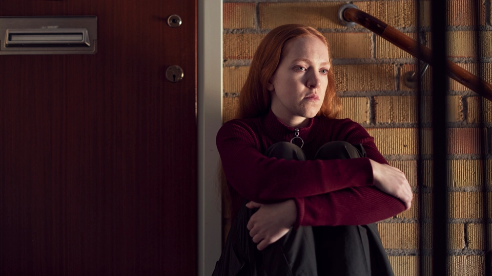 En ung person sitter i ett trapphus och ser nedstämd ut.