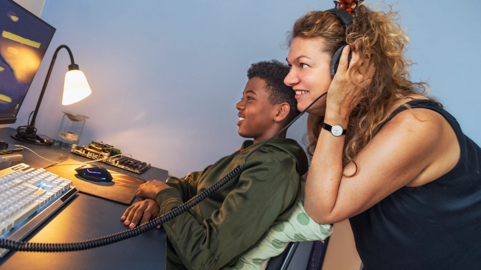 En vuxen kvinna lyssnar i hörlurar tillsammans med sitt barn framför en dator. De ser glada och engagerade ut.