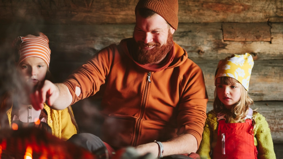 En pappa sitter med sina två döttrar i ett vindskydd. De värmer något att dricka.