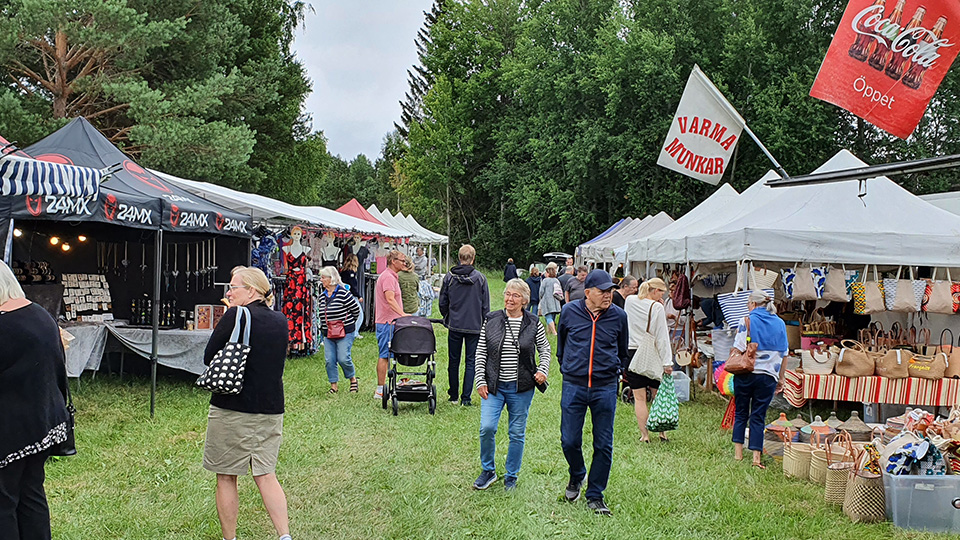 Människor strövar omkring bland marknadsstånd där det säljs blandade varor som väskor, varma munkar och smycken.