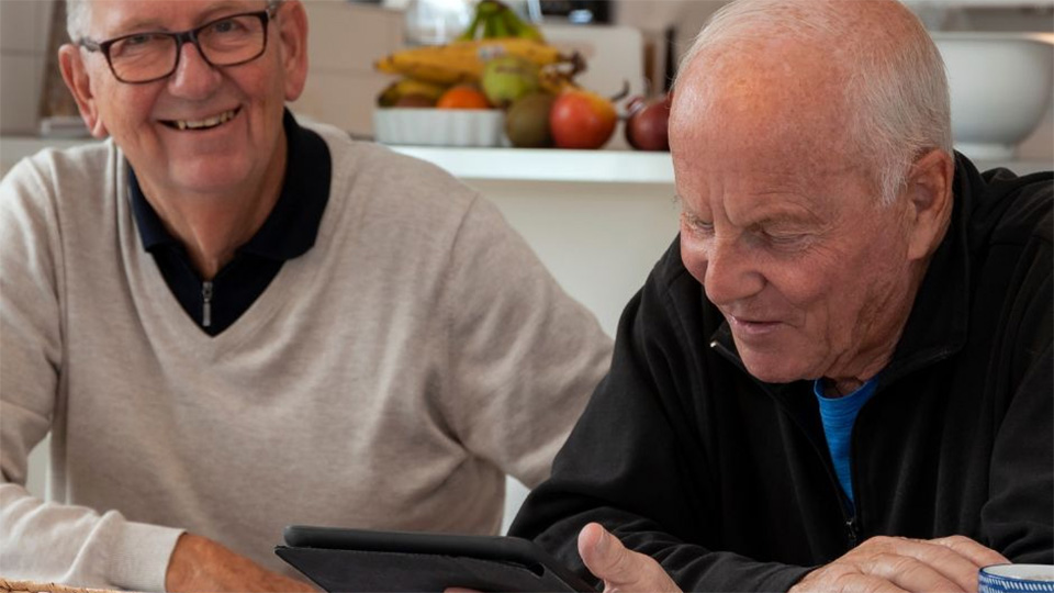 Två äldre herrar. Den ena ler in i kameran den andre ser ned mot en i-Pad han håller i handen.