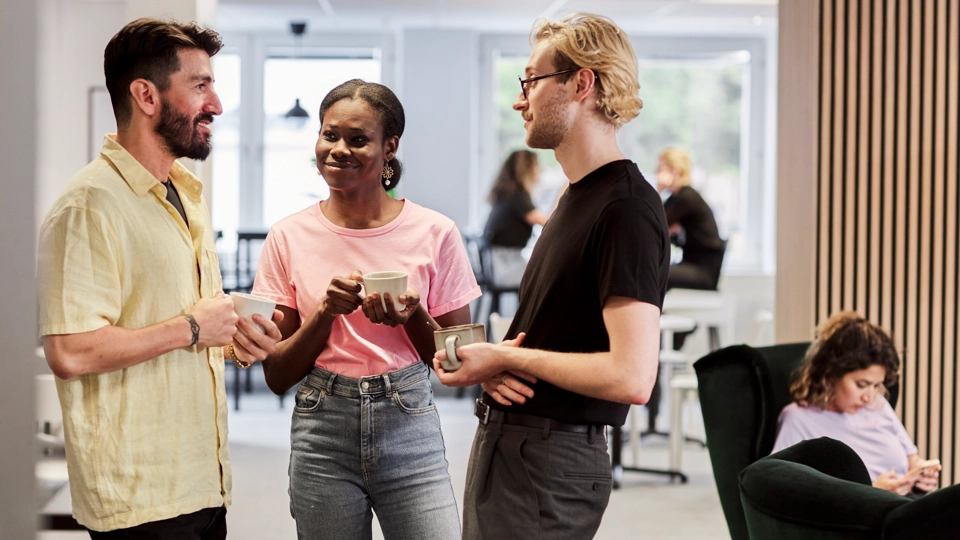 Tre kollegor står och pratar med varandra. En fjärde syns sitta en bit ifrån.
