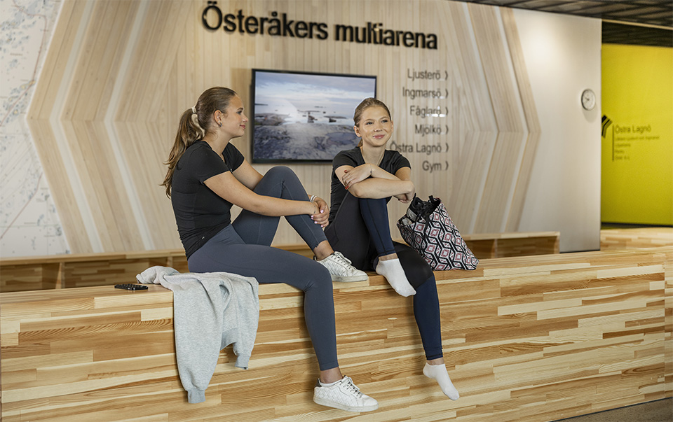 Två unga kvinnor i träningskläder sitter inne i en sporthall.