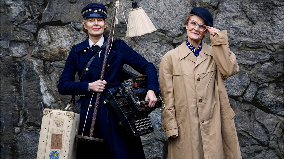 Två kvinnor i kläder från 40-talet. En i mörkblå uniform och den andra i trenchcoat och keps.